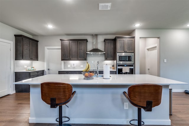 kitchen with appliances with stainless steel finishes, a kitchen breakfast bar, wall chimney exhaust hood, dark brown cabinetry, and a spacious island