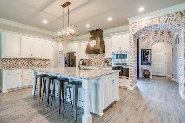 kitchen featuring custom exhaust hood, white cabinetry, appliances with stainless steel finishes, an island with sink, and pendant lighting