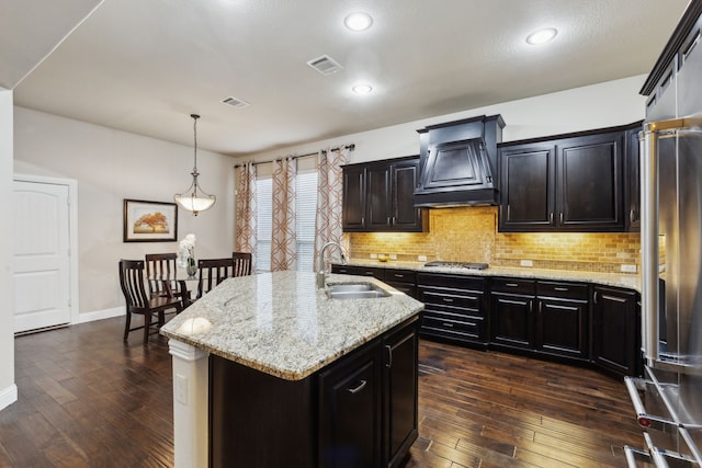 kitchen with premium range hood, sink, hanging light fixtures, dark wood-type flooring, and a center island with sink