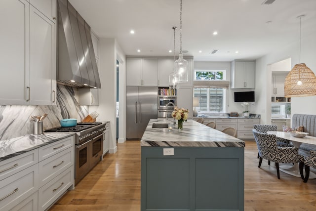 kitchen featuring built in appliances, light hardwood / wood-style floors, an island with sink, decorative light fixtures, and wall chimney exhaust hood