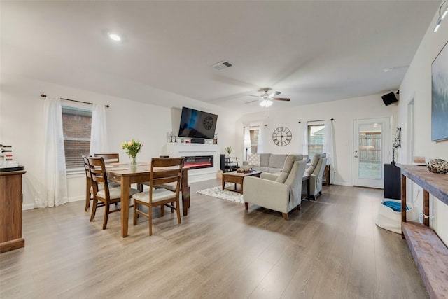 living room featuring light wood finished floors, visible vents, baseboards, a glass covered fireplace, and ceiling fan