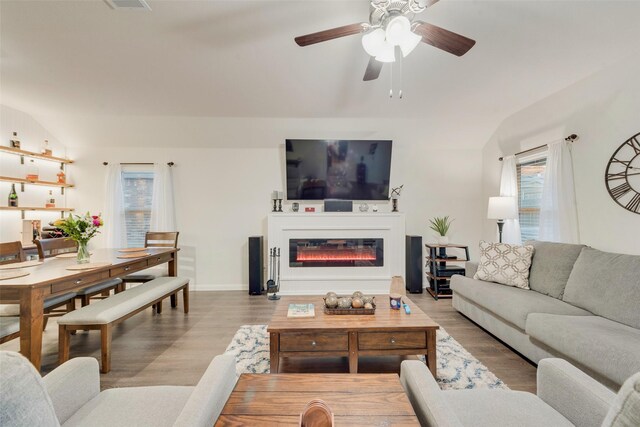 living area featuring light wood finished floors, visible vents, a glass covered fireplace, ceiling fan, and baseboards