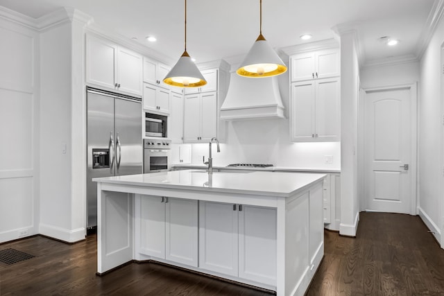 kitchen with premium range hood, built in appliances, hanging light fixtures, a kitchen island with sink, and white cabinets