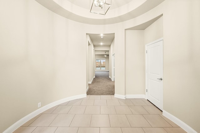 hallway featuring a tray ceiling, light tile patterned floors, baseboards, and an inviting chandelier