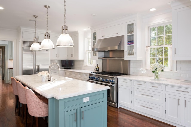 kitchen with wall chimney exhaust hood, high end appliances, white cabinetry, decorative light fixtures, and an island with sink