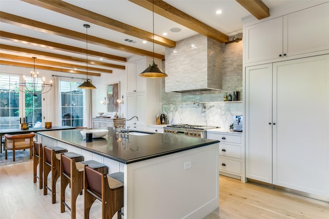 kitchen featuring decorative light fixtures, an island with sink, sink, white cabinets, and wall chimney exhaust hood