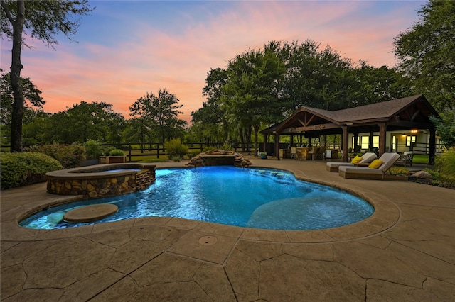 pool at dusk with a patio area, an outdoor structure, pool water feature, and an in ground hot tub