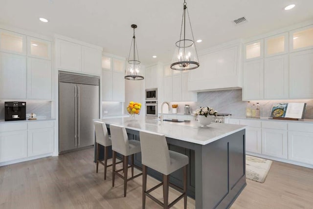 kitchen with built in appliances, sink, white cabinetry, and a center island with sink