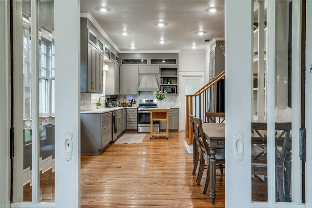 kitchen with custom exhaust hood, light wood-type flooring, ornamental molding, appliances with stainless steel finishes, and backsplash