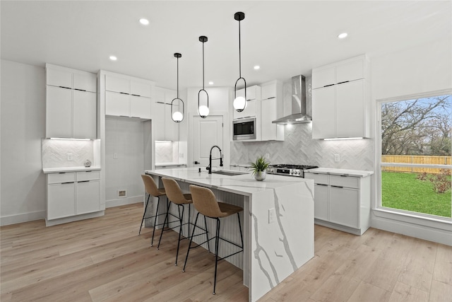 kitchen with sink, stainless steel microwave, wall chimney range hood, a kitchen island with sink, and white cabinets