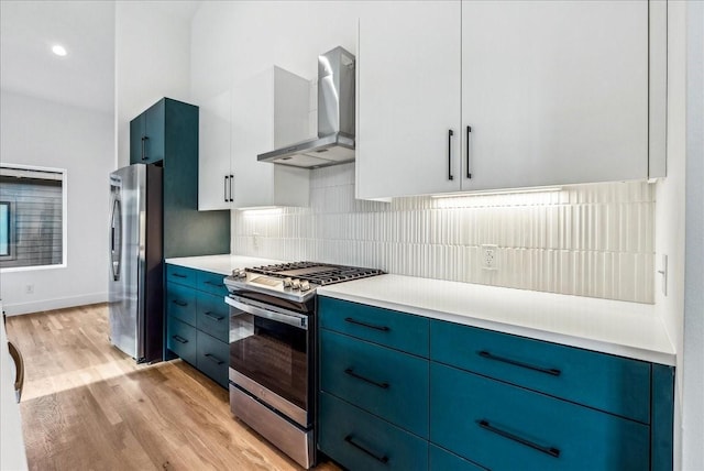 kitchen featuring appliances with stainless steel finishes, blue cabinets, white cabinetry, wall chimney range hood, and light wood-type flooring