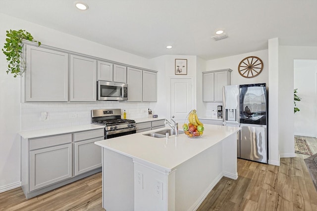 kitchen with appliances with stainless steel finishes, tasteful backsplash, sink, a center island with sink, and light wood-type flooring