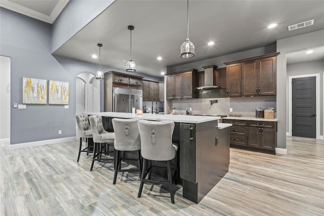 kitchen with pendant lighting, wall chimney range hood, dark brown cabinets, a center island with sink, and stainless steel built in fridge