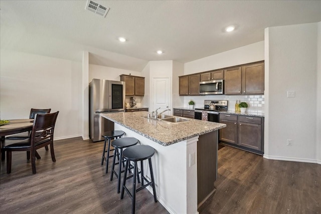 kitchen with dark hardwood / wood-style floors, sink, stainless steel appliances, light stone countertops, and a center island with sink