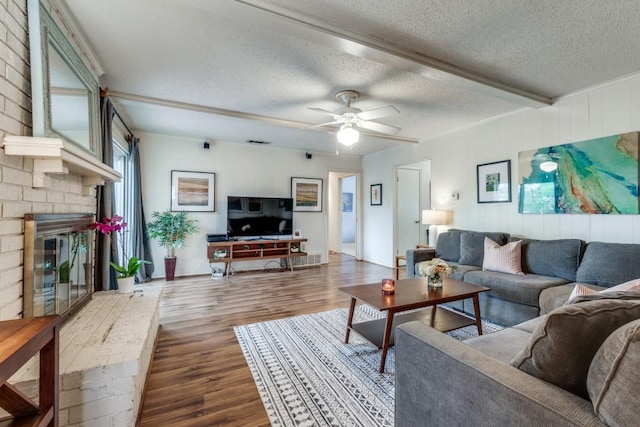 living room with ceiling fan, wood-type flooring, a textured ceiling, a brick fireplace, and beamed ceiling