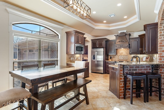 kitchen with premium range hood, appliances with stainless steel finishes, dark brown cabinetry, light stone countertops, and a raised ceiling