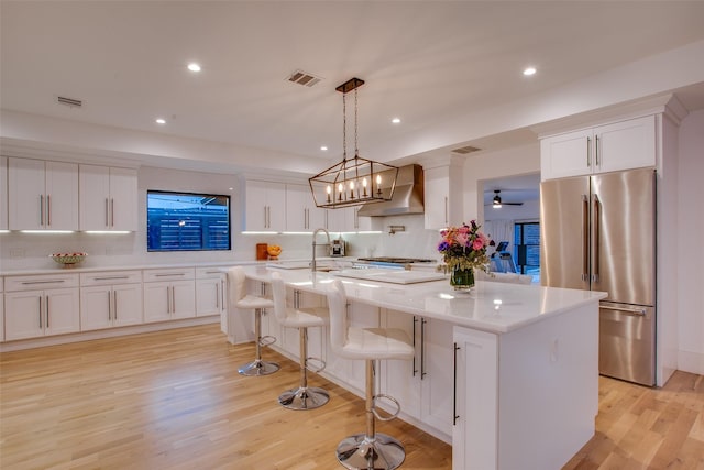 kitchen with an island with sink, high quality fridge, wall chimney range hood, and white cabinets