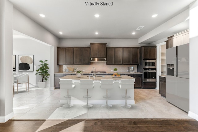 kitchen with backsplash, stainless steel appliances, a kitchen breakfast bar, dark brown cabinetry, and a center island with sink