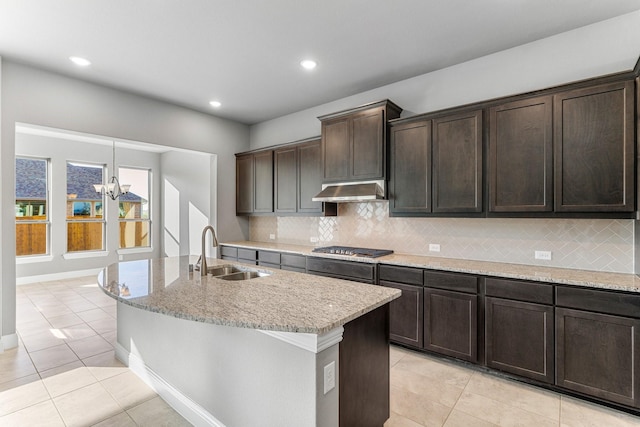 kitchen with sink, tasteful backsplash, light stone counters, dark brown cabinets, and a kitchen island with sink