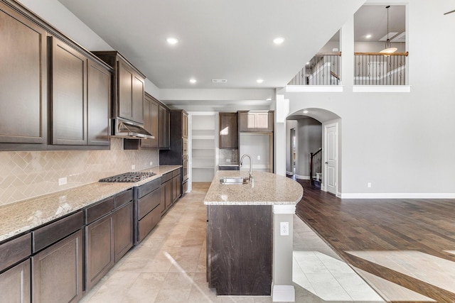 kitchen with sink, light stone counters, dark brown cabinets, a kitchen island with sink, and backsplash