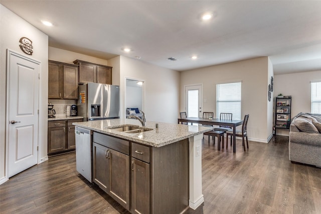 kitchen with appliances with stainless steel finishes, an island with sink, sink, light stone counters, and dark wood-type flooring