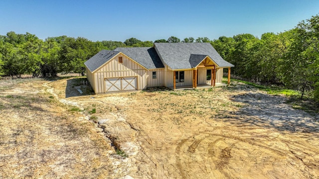 property in mid-construction with board and batten siding, a porch, a garage, dirt driveway, and roof with shingles