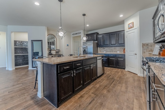 kitchen featuring appliances with stainless steel finishes, hardwood / wood-style floors, pendant lighting, light stone countertops, and a center island with sink