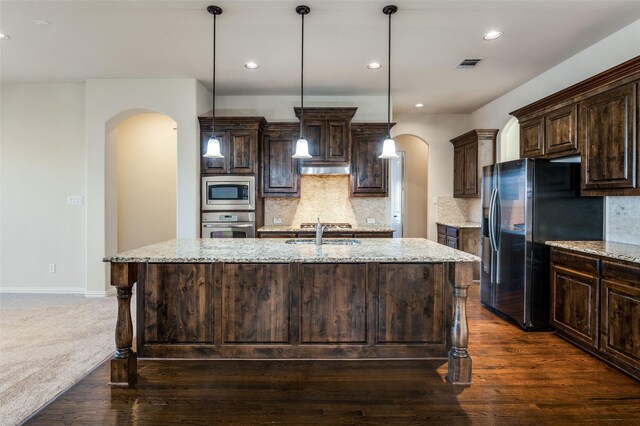 kitchen with appliances with stainless steel finishes, light stone counters, tasteful backsplash, a center island with sink, and decorative light fixtures