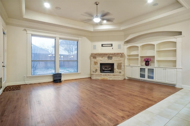 unfurnished living room with a stone fireplace, ornamental molding, a tray ceiling, ceiling fan, and hardwood / wood-style floors