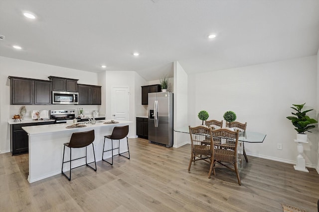 kitchen with light hardwood / wood-style flooring, appliances with stainless steel finishes, dark brown cabinets, an island with sink, and a kitchen bar