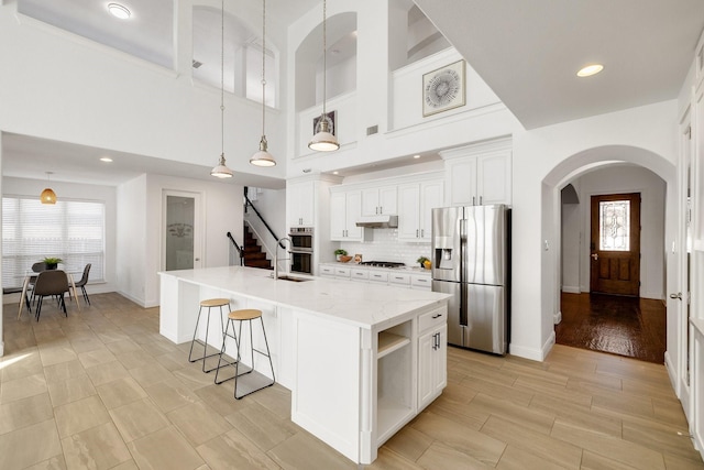 kitchen with white cabinetry, appliances with stainless steel finishes, pendant lighting, light stone countertops, and a kitchen island with sink