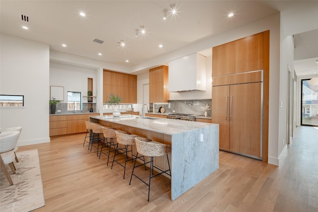 kitchen featuring wall chimney exhaust hood, light stone counters, light hardwood / wood-style flooring, a large island with sink, and a kitchen breakfast bar