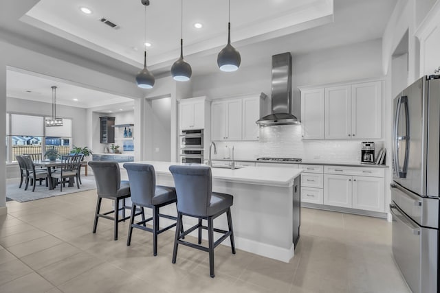 kitchen with pendant lighting, stainless steel appliances, a tray ceiling, a center island with sink, and wall chimney exhaust hood