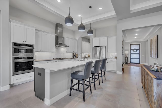 kitchen with a raised ceiling, white cabinetry, a kitchen island with sink, and wall chimney exhaust hood