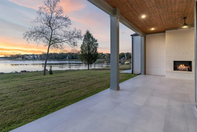 patio terrace at dusk with an outdoor brick fireplace, a water view, ceiling fan, and a lawn