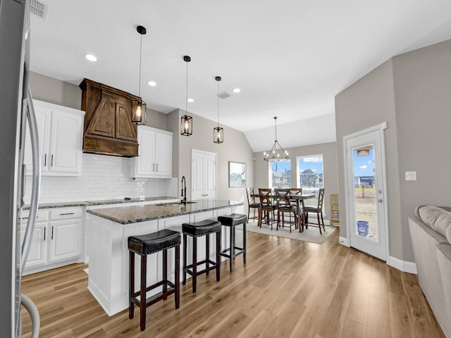 kitchen featuring stainless steel fridge, white cabinetry, dark stone countertops, custom range hood, and an island with sink