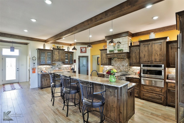 kitchen with light stone counters, stainless steel oven, dark brown cabinetry, and a kitchen island with sink