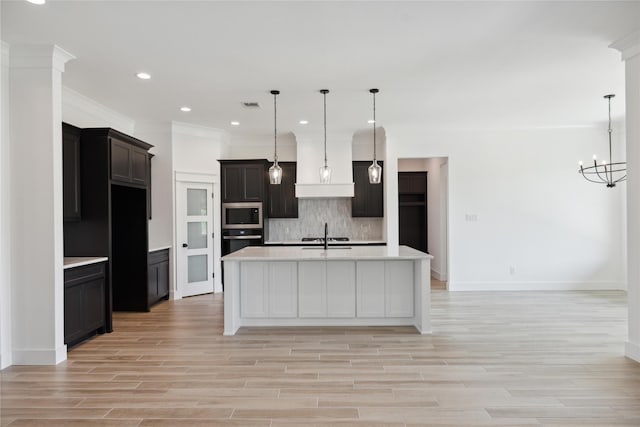 kitchen featuring a kitchen island with sink, backsplash, stainless steel appliances, decorative light fixtures, and light wood-type flooring