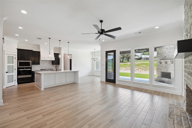 unfurnished living room featuring sink, ceiling fan with notable chandelier, ornamental molding, a stone fireplace, and light wood-type flooring