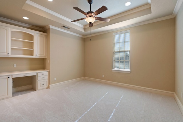 unfurnished office with built in desk, ornamental molding, light colored carpet, ceiling fan, and a raised ceiling