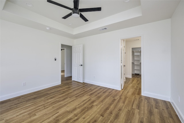 unfurnished bedroom with hardwood / wood-style floors, a walk in closet, a raised ceiling, and ceiling fan