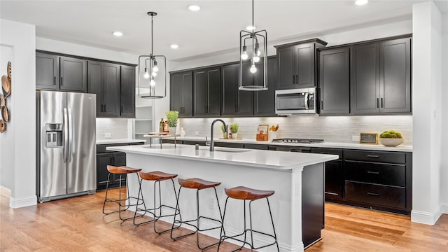 kitchen with sink, a center island with sink, light wood-type flooring, pendant lighting, and stainless steel appliances