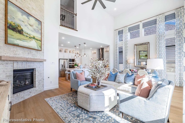 living room featuring ceiling fan, a brick fireplace, a towering ceiling, and light wood-type flooring