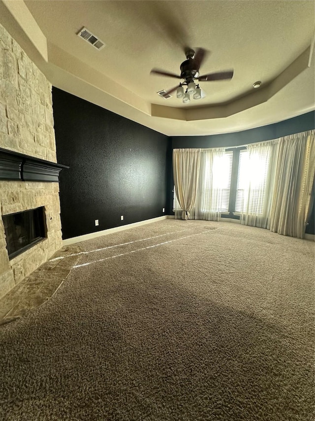 unfurnished living room featuring a stone fireplace, a textured ceiling, a tray ceiling, carpet floors, and ceiling fan