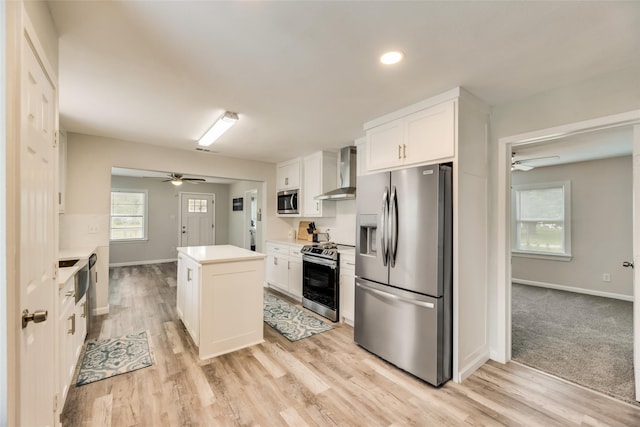 kitchen featuring wall chimney range hood, appliances with stainless steel finishes, white cabinetry, a kitchen island, and light wood-type flooring