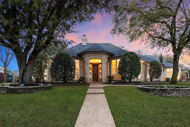 french country home with a shingled roof, brick siding, a lawn, and a chimney