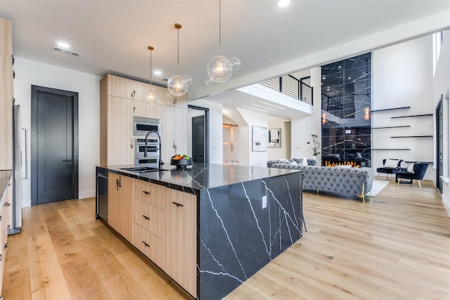 kitchen featuring appliances with stainless steel finishes, light brown cabinets, hanging light fixtures, a center island with sink, and light wood-type flooring