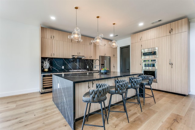 kitchen with dark stone countertops, hanging light fixtures, stainless steel appliances, a center island with sink, and light brown cabinetry