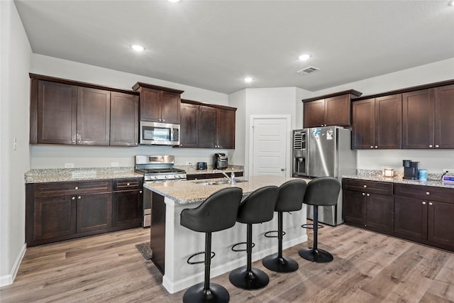 kitchen with dark brown cabinetry, light stone counters, stainless steel appliances, and light wood-type flooring