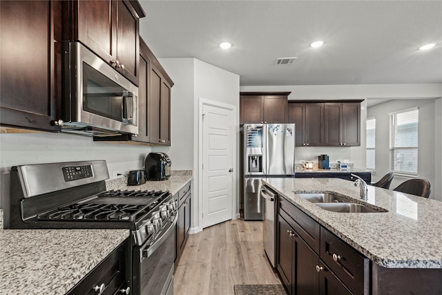 kitchen with sink, appliances with stainless steel finishes, a kitchen island with sink, light hardwood / wood-style floors, and light stone countertops